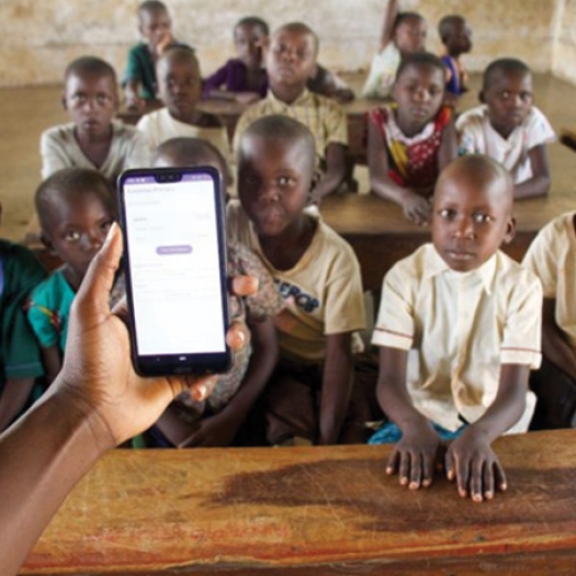 A hand extends forward holding a smartphone with the Skizaa app on the screen in front of a classroom full of elementary school children in Uganda's Mayuge district.