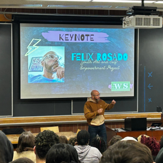 Felix Rosado, the program coordinator for Healing Futures with the Youth Art & Self-empowerment Project (YASP) and an adjunct professor at Chestnut Hill College, delivers a keynote to an audience of high school students in front of a projector screen with his headshot and name on it in a lecture hall.