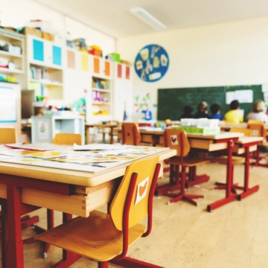 A cluster of wooden student desks in a bright, sun-filled elementary school classroom. In the background, out of focus, appears to be a reading circle. Credit: Adobe Stock.