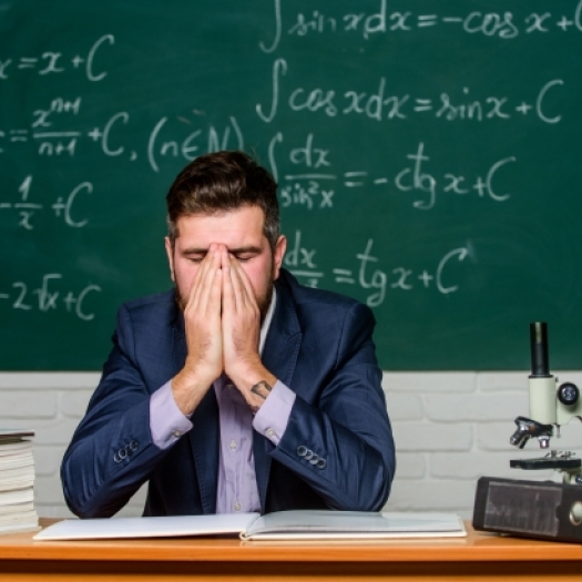 A young teacher in a suit sits at a desk in front of a chalkboard covered with mathematical equations. He has his hands covering his face in a gesture of exhaustion or frustration. The desk has thick stacks of books, papers, a binder and a microscope. He looks tired and overwhelmed.