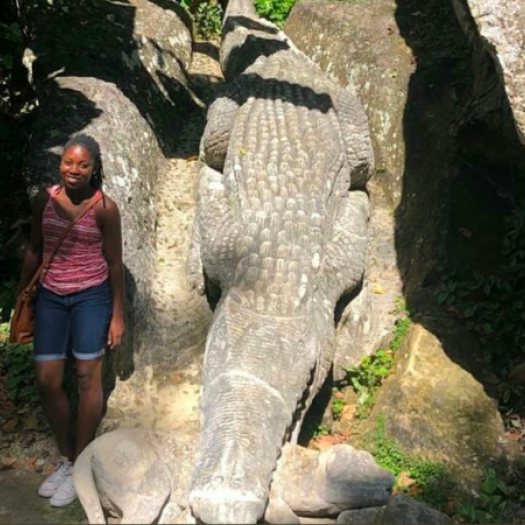 Angela Crumdy stands beside a large statue of an alligator biting another animal, surrounded by trees and rocks in Guantánamo, Cuba.