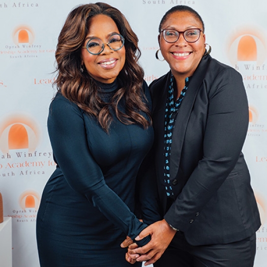 Oprah Winfrey stands in a dark blue dress, clasping hands with Penn GSE Assistant Adjunct Professor Charlotte Jacobs on the right, who is wearing a black suit over a black-and-blue button-down shirt. The two stand in front of a backdrop of the Oprah Winfrey Leadership Academy for Girls logo behind them.