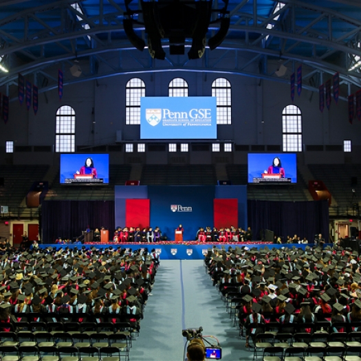 Penn GSE University of Pennsylvania, with graduates seated in the Palestra under banners, facing a stage where a Angela Duckworth is visible on two large screens.