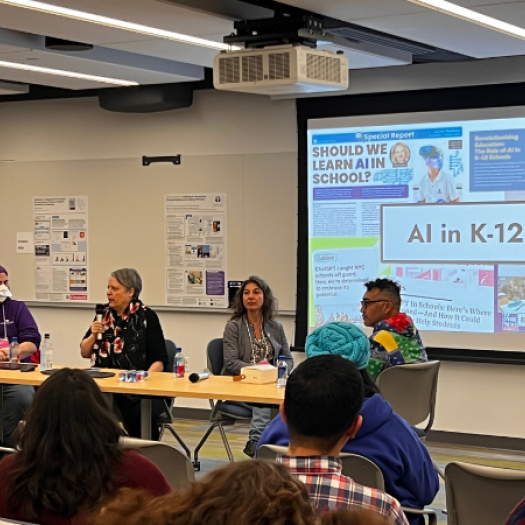 Four people sit in a row at a long table in front of an audience with a slide that says “AI in K–12” on a screen behind them.