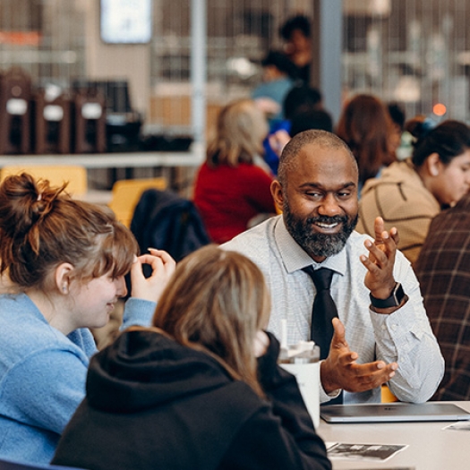 A teacher – a Black man with a trimmed beard in a white shirt and tie – sits facing the camera. He is at a table during a breakout session. He is smiling animatedly and speaking with a small, diverse group of students. Similar groups working at other tables are slightly out of focus in the background.