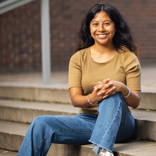 Penn GSE student Meresa García sits outside on cement steps in front of a brick wall on the University of Pennsylvania campus wearing a tan t-shirt and blue jeans, smiling at the camera