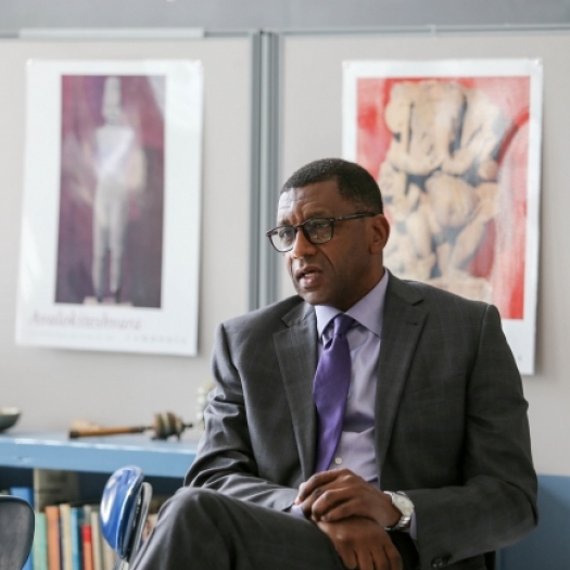 Dr. Howard Stevenson is seated in front of a high school classroom, facing students who are out of focus in the foreground. He appears relaxed and casual, resting his hands in his lap. He is wearing a grey suit with a purple tie and is listening to one of the students off-camera who is sharing an experience.