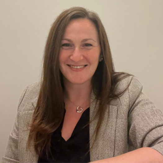 A headshot portrait of Lara wearing a herringbone blazer and smiling against a neutral background. 