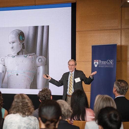 Dr. James Lester, Distinguished University Professor of Computer Science and Director of the Center for Educational Informatics at North Carolina State University, stands with his arms spread in front of a seated crowd while delivering the keynote at the launch of Penn GSE's McGraw Center for Educational Leadership. A white robot can be seen on the screen behind him. The topic of the talk was AI & Education.