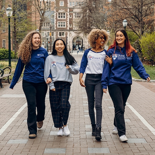 Four Penn GSE students wearing Penn GSE sweatshirts in blue, gray, or white walk down a path on the Penn campus. They are smiling and laughing.