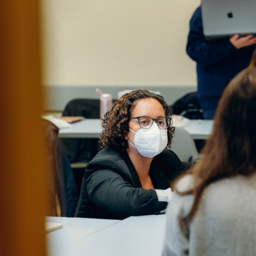 A photo in a classroom. Professor Sade Bonilla is leaning down at desk level to talk with two of her seated students whose backs are to the camera. They are all deep in conversation.