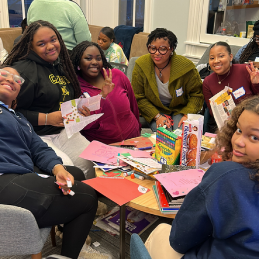 A group of teenage high school students hanging out during a recent Black Girls Literacies Project meeting. Everyone is casually dressed and comfortable. They’re seated around a coffee table piled high with school boxes, art supplies and snacks. They’re in the middle of doing a project. Everyone is happy and smiling, and several of the students are making the peace sign with their hands. It looks fun and friendly.