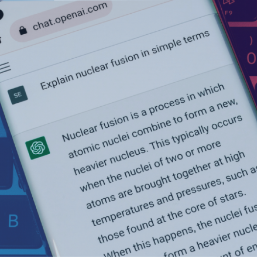 A smartphone is held in the center of the image with the ChatGPT website open on its browser. The phone is above a computer keyboard. You can see that the person has input “explain nuclear fusion in simple terms” as their prompt and the AI is generating an answer in return, illustrating how the program works for end-users.