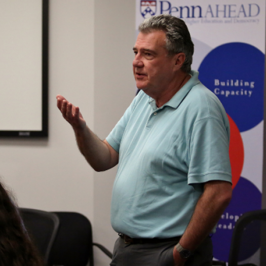 Alan Ruby teaching a class. He’s standing in a classroom, talking, while facing tables of students listening and taking notes. A banner behind him promotes the Penn AHEAD program, where Ruby is a senior fellow.