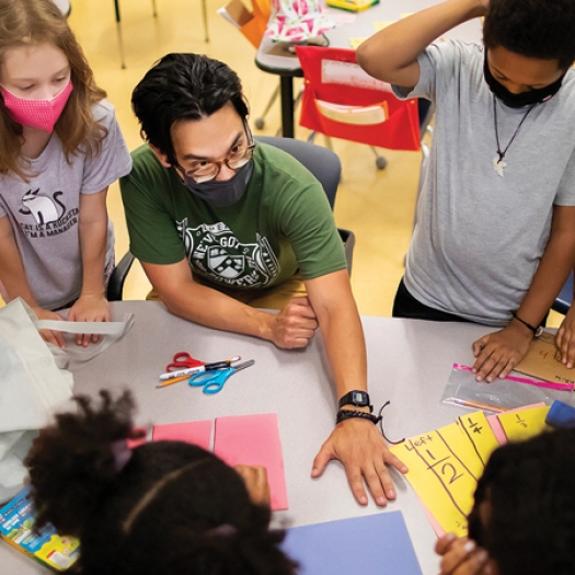 A seated teacher, wearing a face mask and glasses, is surrounded by several masked elementary students and reaches across a table to engage with a student's paper.