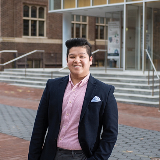 Smiling young man in red-striped shirt and jacket standing outside on Penn’s campus. 