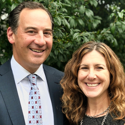 A man in a suit and tie and a woman in a black blouse smiling while standing outdoors