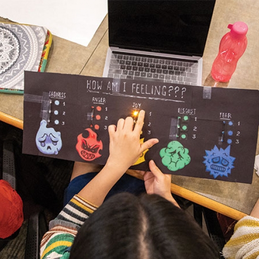 Aerial view of a student holding a colorful chart with a list of emotions and a light shining in the middle underneath the word “Joy.”