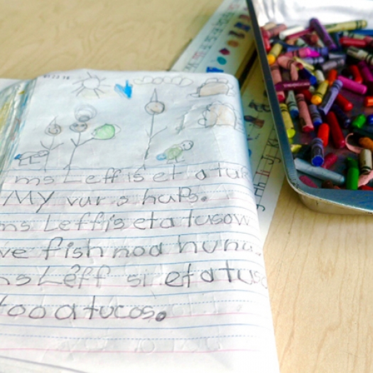 A folded composition book with children’s writing and drawings lies next to a tray of crayons 