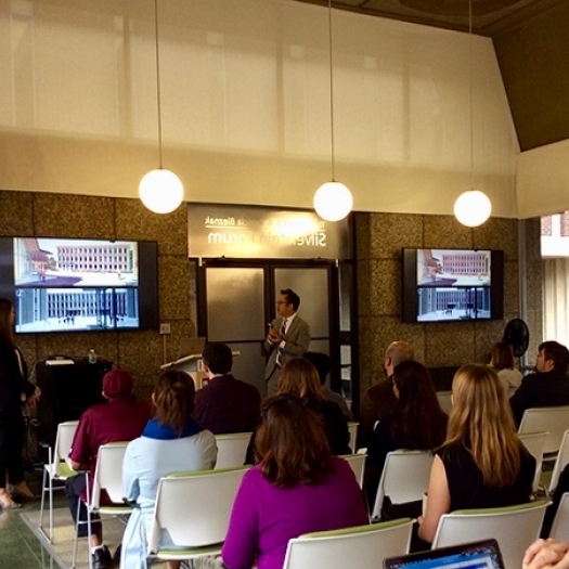 Man speaking in front of audience, with images of a building on screens behind him