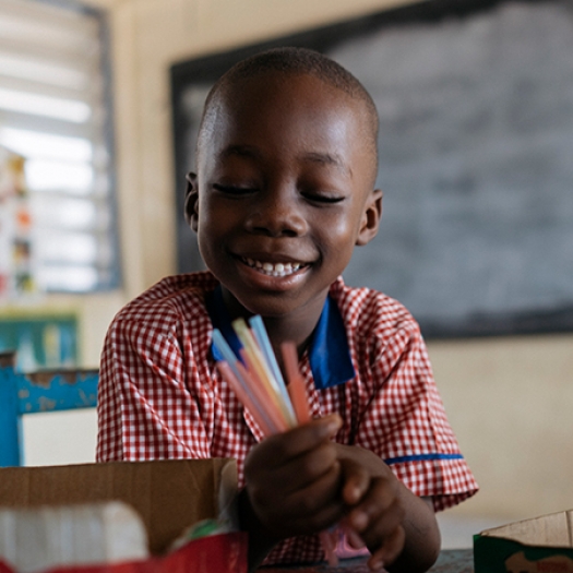 A child in a classroom in Ghana smiles while holding a handful of pens