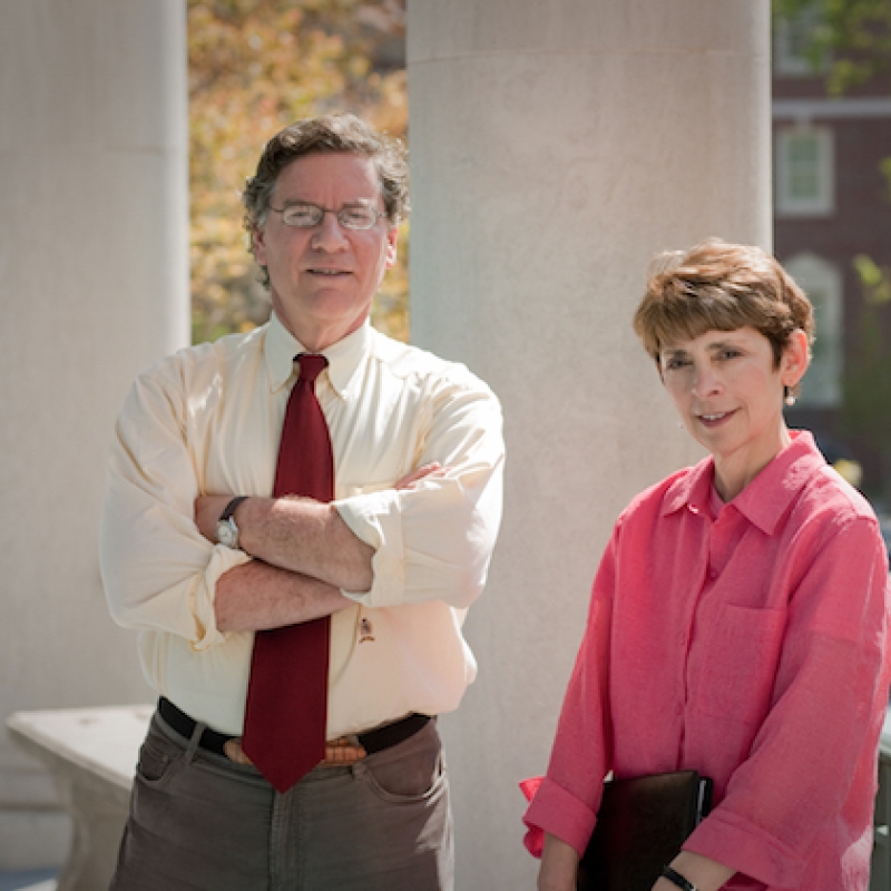 Man with glasses standing and woman with short hair sitting outside.