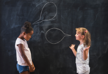 Two elementary school students speak and gesture to each other in a contentious conversation in front of a chalkboard with chalk speech bubbles drawn over each.