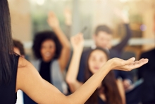 A woman instructor’s arm outstretched in the “I don’t know” gesture while people seated in front of her raise their hands with questions.