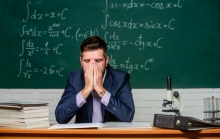 A young teacher in a suit sits at a desk in front of a chalkboard covered with mathematical equations. He has his hands covering his face in a gesture of exhaustion or frustration. The desk has thick stacks of books, papers, a binder and a microscope. He looks tired and overwhelmed.
