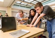 Three students gathered around a table, one pointing at a small appliance