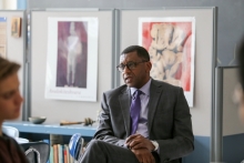 Dr. Howard Stevenson is seated in front of a high school classroom, facing students who are out of focus in the foreground. He appears relaxed and casual, resting his hands in his lap. He is wearing a grey suit with a purple tie and is listening to one of the students off-camera who is sharing an experience.