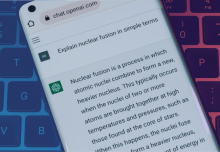 A smartphone is held in the center of the image with the ChatGPT website open on its browser. The phone is above a computer keyboard. You can see that the person has input “explain nuclear fusion in simple terms” as their prompt and the AI is generating an answer in return, illustrating how the program works for end-users.