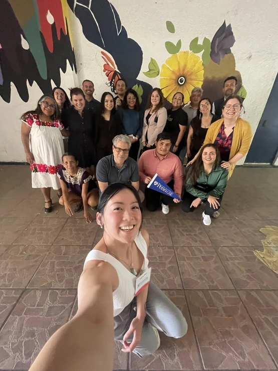 A group of people stand in front of a mural while a woman in the foreground takes a selfie with everyone behind her