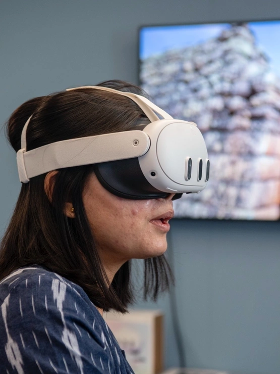 a woman with straight hair wears VR goggles in front of a wall with a large monitor mounted