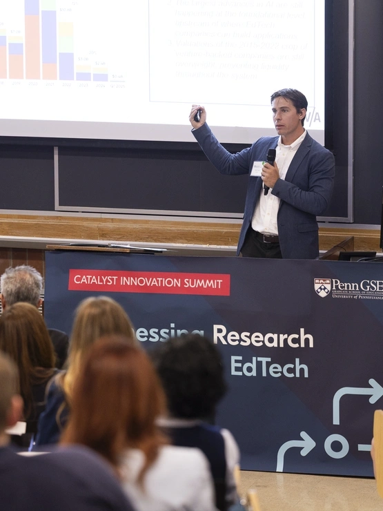 Matt stands in front of an audience wielding a clicker and pointing at a screen that is displaying a large graph