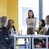 Women seated around a table having a conversation, with a woman standing over them arms crossed and observing.