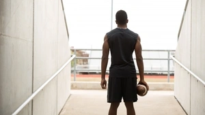 A teen in gym clothes holding a football stands in the entrance to a stadium looking out onto the field.