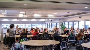 A group of more than 30 teacher education researchers gathered around large round tables in a conference room listening to an attendee speaking into a microphone while the presenters stand at the front.