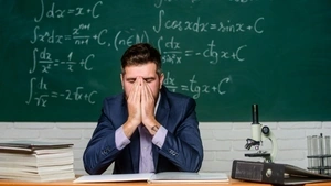 A young teacher in a suit sits at a desk in front of a chalkboard covered with mathematical equations. He has his hands covering his face in a gesture of exhaustion or frustration. The desk has thick stacks of books, papers, a binder and a microscope. He looks tired and overwhelmed.
