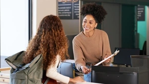 A smiling counselor leans over a counter with a clipboard while helping a teen with a backpack.