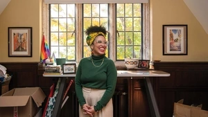 Krista Cortes stands, hands folded in front of her, laughing in the middle of her office surrounded by decorations showcasing Latin American culture.