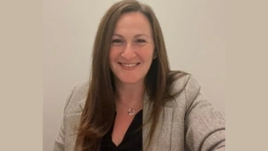 A headshot portrait of Lara wearing a herringbone blazer and smiling against a neutral background. 