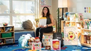 Sarah sits in her classroom holding a packet of letters and drawings from students and surrounded by puppets, books, pillows, and costumes that she uses in her class.