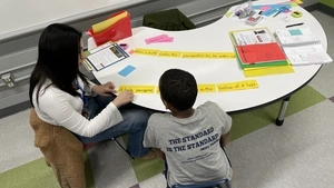 A Harrison Elementary student and his Penn tutor are working through the sentence "A baby porcupine... in the hollow of a tree" while seated at a kidney-shaped table in a classroom.