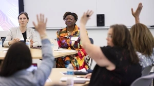 A group of three people have their hands up and visible through their arms are two women speaking at the front of the room. 