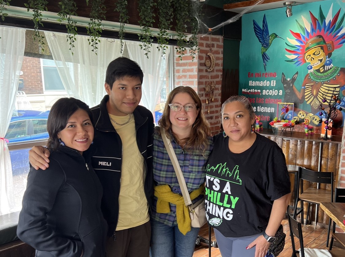 Four people stand together with their arms around one another in a restaurant, smiling at the camera. Behind them is a window on one wall and a mural of an Aztec god on another.