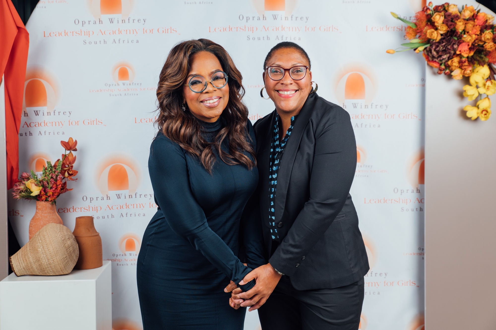 Oprah Winfrey stands in a dark blue dress, clasping hands with Penn GSE Assistant Adjunct Professor Charlotte Jacobs on the right, who is wearing a black suit over a black-and-blue button-down shirt. The two stand in front of a backdrop of the Oprah Winfrey Leadership Academy for Girls logo behind them.
