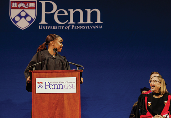 Abbott Elementary creator Quinta Brunson stands at a podium sharing a laugh with Penn GSE Dean Pam Grossman, seated to her right.