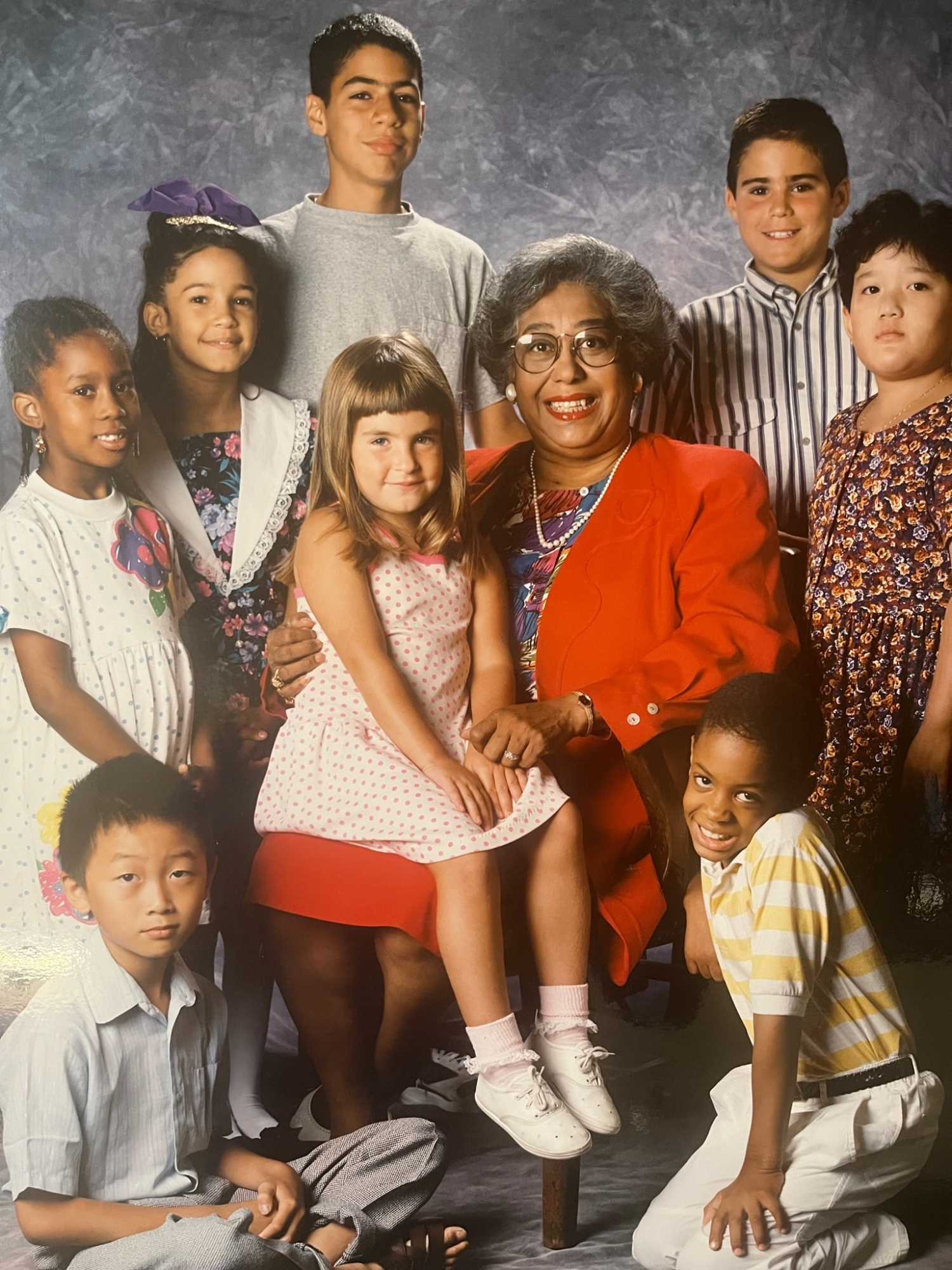 Dr. Constance E. Clayton sits in a chair wearing a red skirt and blazer over a floral shirt, surrounded by a multiracial group of children from the Philadelphia School District, during her term as Philadelphia