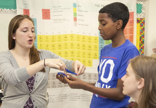A teacher puts blue rubber bands in a student’s hand standing in front of a giant poster of the periodic table of elements.